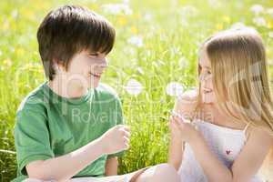 Two young children sitting outdoors holding dandelion heads smil