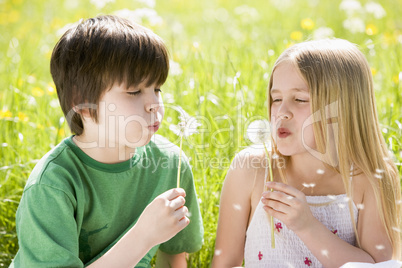 Two young children sitting outdoors blowing dandelion heads smil