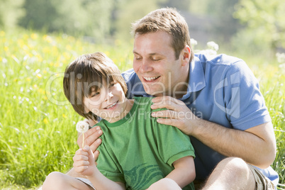 Father and son sitting outdoors with dandelion head smiling