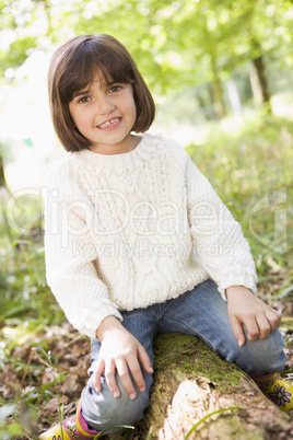 Young girl outdoors in woods sitting on log smiling