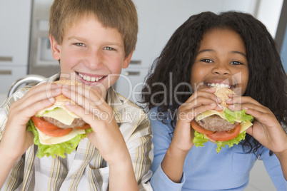 Two young children in kitchen eating cheeseburgers smiling