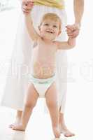 Mother helping baby walk indoors smiling