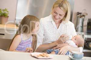 Mother feeding baby in kitchen with daughter eating cookies and