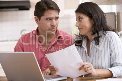 Couple in kitchen with paperwork using laptop looking unhappy