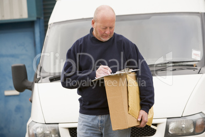 Delivery person standing with van writing in clipboard holding box
