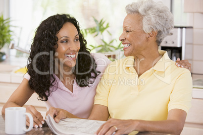 Two women in kitchen with newspaper and coffee smiling