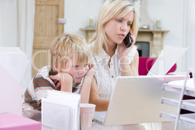 Woman using telephone in home office with laptop while young boy