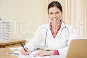 Doctor with laptop writing in doctor's office smiling