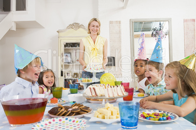 Young children at party sitting at table with mother carrying cake
