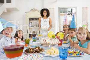 Young children at party sitting at table with mother carrying cake