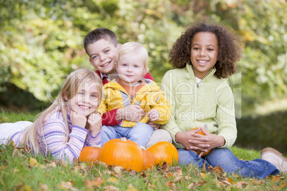 Three young friends with baby sitting on grass with pumpkins smi