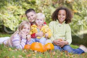 Three young friends with baby sitting on grass with pumpkins smi