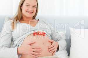 Blond pregnant woman with mom letters on her belly lying on a be