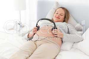 Smiling future mom putting headphones on her belly lying on a be
