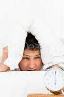 Tired man with head under the pillow looking at his clock