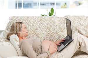 Smiling pregnant woman using her laptop lying on the sofa