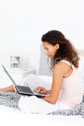 Serious hispanic woman working on her laptop sitting on her bed