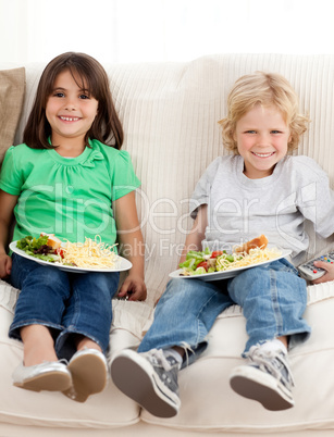 Happy brother and sister watching television while eating pasta