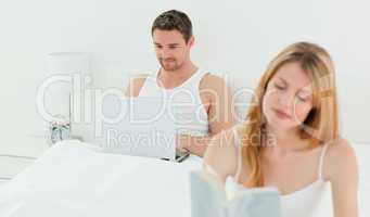 Man is on his laptop while his wife is reading a book