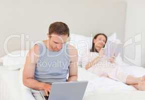 Manworking on his laptop while his wife is reading a book on the