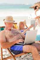 Man working on his laptop while his wife is reading at the beach