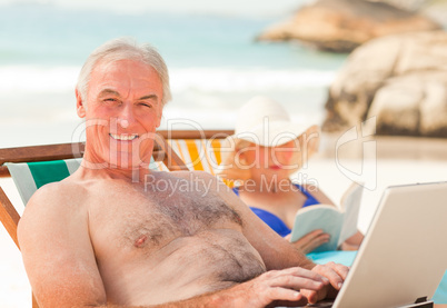 Man working on his laptop while his wife is reading at the beach