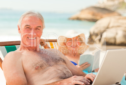 Man working on his laptop while his wife is reading at the beach