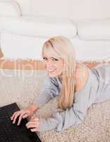 Attractive young blond woman relaxing on laptop lying on a carpe