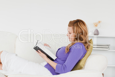 Pretty red-haired woman reading a book while lying on a sofa