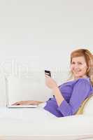 Smiling red-haired woman using a laptop and a phone while lying