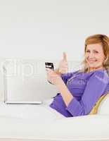 Happy red-haired woman using a laptop and a phone while lying on