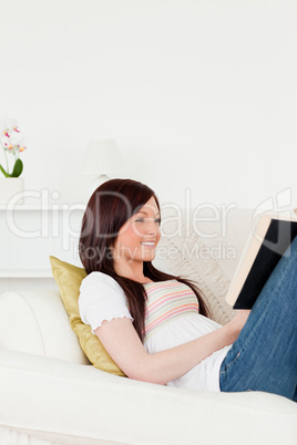 Charming red-haired woman reading a book while lying on a sofa