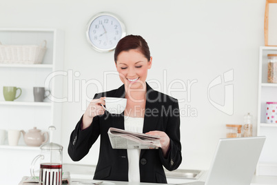 Pretty red-haired woman in suit reading the newspaper while havi