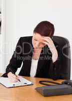 Depressed beautiful red-haired woman in suit writing on a notepa