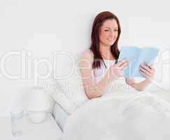 Pretty red-haired woman reading a book while sitting on her bed