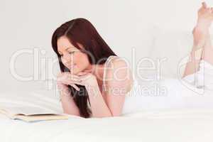 Beautiful red-haired woman reading a book while lying on her bed