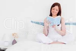 Cute red-haired woman reading a book while sitting on her bed
