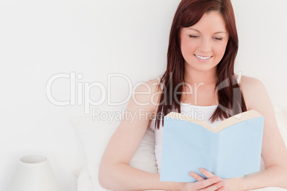 Pretty red-haired woman reading a book while sitting on her bed