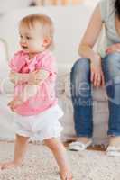 Baby standing on a carpet while her mother is sitting on a sofa