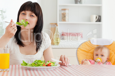 Attractive brunette woman eating a salad next to her baby while