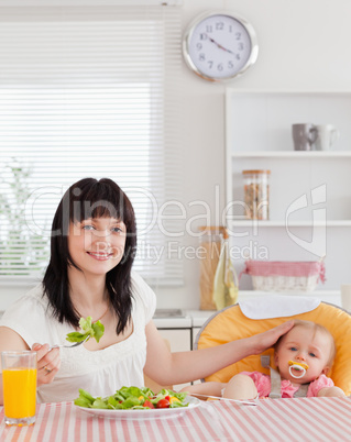 Pretty brunette woman eating a salad next to her baby while sitt