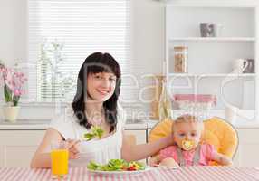 Charming brunette woman eating a salad next to her baby while si