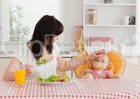 Gorgeous brunette woman eating a salad next to her baby while si