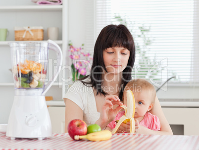 Cute brunette woman pealing a banana while holding her baby on h