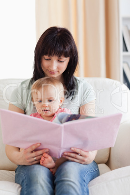 Pretty woman holding her baby and a book in her arms while sitti