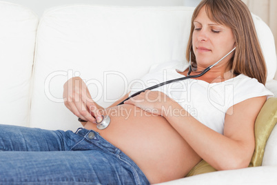 Charming pregnant woman using a stethoscope while lying on a sof