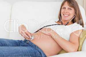 Beautiful pregnant woman using a stethoscope while lying on a so