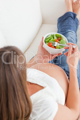 Good looking pregnant woman eating a salad while lying on a sofa