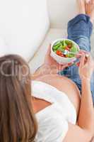 Good looking pregnant woman eating a salad while lying on a sofa