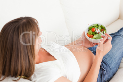 Charming pregnant woman eating a salad while lying on a sofa